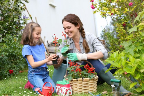 A woman and a young girl are gardening together in a lush garden, surrounded by vibrant green foliage, flowering plants, and a well-maintained lawn in Pimlico. The woman, dressed casually with green gardening gloves, is holding a small potted plant with red flowers, smiling as she assists the girl. The girl, wearing a blue t-shirt and denim shorts, is carefully holding the plant, engaging in a gardening activity. Nearby, there is a wicker basket filled with gardening tools and supplies, including a red watering can and a small patterned container. The garden features a mix of leafy shrubs, flowering plants, and a paved pathway, with sunlight filtering through the leaves, creating a bright outdoor environment ideal for gardening and outdoor maintenance services provided by Garden Maintenance Pimlico. The scene emphasizes a nurturing and natural setting, illustrating typical backyard gardening efforts in an urban outdoor space.
