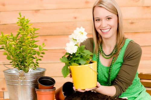 A woman with light brown hair smiling outdoors in a garden setting, seated in front of a wooden fence, holding a yellow flower pot containing a flowering plant with white blooms and green foliage. To her left, there is a large potted shrub with bright green leaves in a silver metal container, and nearby are several empty plant pots of varying sizes and colors, including black and terracotta. The garden features a well-maintained lawn with lush, dense grass, and the scene suggests ongoing or recent gardening work, with natural lighting indicating a bright, clear day. The overall environment appears neat and inviting, suitable for gardening and outdoor maintenance activities typical of a residential garden in Pimlico or nearby London areas.
