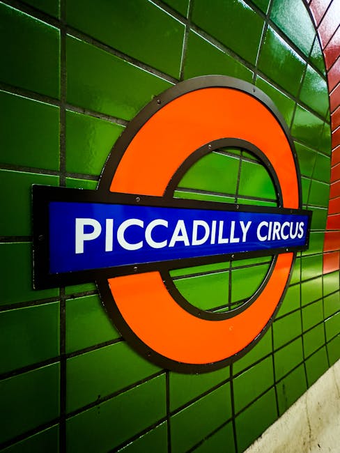 A close-up view of the iconic Piccadilly Circus London Underground station sign mounted on a vibrant green tiled wall. The sign features a bold orange circle with a black outline, intersected by a blu